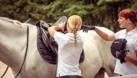 Mentor saddling a white horse for her student, who is pointing at the saddle and holding a helmet. Trees in the background.